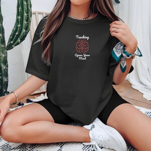 May include: Black t-shirt with the text "Teaching Opens Your Mind" and a pink brain graphic. The shirt is a crew neck and appears to be made of a soft material. The shirt is worn with black shorts and white sneakers.