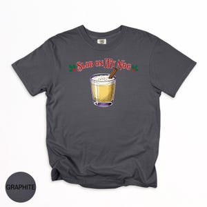 May include: Graphite-colored t-shirt featuring a graphic of a festive eggnog drink with a cinnamon stick. The text "Slob on my Nog" is arched above the drink, with holly accents. The shirt is a comfortable, casual style.