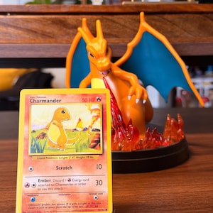 Puede incluir: Una figura de Charizard con alas azules y cuerpo naranja está detrás de una carta coleccionable de Pokémon. La carta presenta una ilustración de Charmander con el texto "Charmander" y "50 HP". La carta también enumera la longitud y el peso de la criatura.