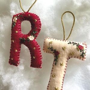 May include: Two felt letter ornaments, "R" in red and "T" in cream, with gold string hangers. The letters are decorated with embroidered flowers and gold star accents, perfect for holiday decor or personalized gifts.