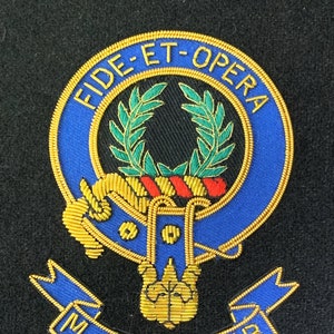 Op de afbeelding: Een geborduurd wapenschild in blauw en goud met de tekst "Fide Et Opera" en "MacArthur" op een zwarte achtergrond.