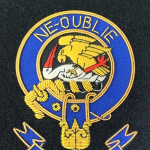 Puede incluir: Un escudo bordado en azul y dorado con el texto "NE-OUBLIE GRAHAM". El escudo presenta un pájaro con un pico rojo y un pájaro amarillo con un ala roja.