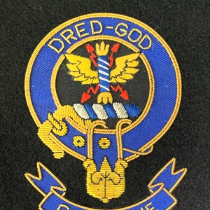 Könnte beinhalten: Ein gesticktes Wappen in Blau und Gold mit dem Text "DRED-GOD CARNEGIE" außen herum. Das Wappen zeigt einen stilisierten Adler mit einer Krone auf dem Kopf und einem Blitz in seinen Krallen.