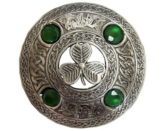 Puede incluir: Un broche celta plateado con un diseño circular. El broche presenta un trébol central y cuatro acentos de piedras preciosas verdes. Intricados nudos y figuras de animales adornan el anillo exterior, creando una apariencia detallada y ornamentada.