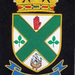 May include: A gold embroidered crest featuring a green cross, a red hand, and a ship. The crest is topped with a gold crown and has a blue ribbon with the word "HUGHES" on it.