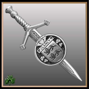 May include: A silver-toned kilt pin featuring a sword with a shield. The shield has a crest with two lions and the text "Clancy" on it. The shield is surrounded by shamrocks.