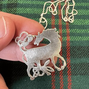 May include: A silver necklace with a pendant featuring a detailed silhouette of a unicorn and a smaller animal, possibly a dog or a cat, standing beneath it.