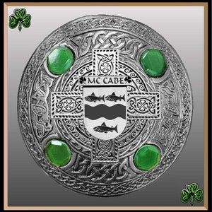 Könnte beinhalten: Eine silberne Kiltnadel mit keltischen Knoten, grünen Edelsteinen und dem Text "MC CABE" in schwarzen Buchstaben.