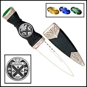 May include: A black and silver Scottish dirk with a green and silver handle. The handle features a silver crest with the text "Hughes" and a Celtic knot. The dirk comes with a black leather scabbard with silver accents.