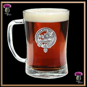 May include: A clear glass beer mug with a handle, filled with a dark amber coloured beer. A silver clan crest with a boar's head and the text "NUNQUAM OBLIVISCAR" is attached to the mug.