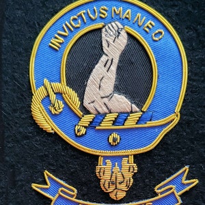 May include: A blue and gold embroidered crest with a raised arm and the text "INVICTUS MANEO" above a blue ribbon with the text "ARMSTRONG".