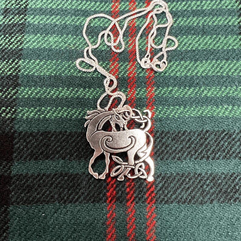 May include: A silver Celtic knot pendant necklace featuring a stylized deer design. The pendant is on a green and red plaid fabric background.