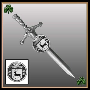 May include: A silver-colored kilt pin in the shape of a sword with a Celtic knot design. The sword has a round shield at the base with a black and white image of a stag and the text "O'Connor".