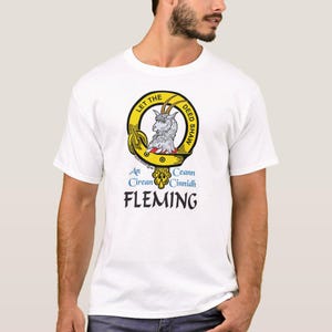 May include: White t-shirt with a yellow circular emblem. The emblem features a goat illustration and the text "LET THE DEED SHAW". Below the emblem are the words "An Cirean Cinnidh" and "FLEMING" in blue. The shirt has short sleeves.