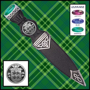 May include: A silver Clan Clancy crest with a green shamrock and a green emerald gemstone on a black leather sgian dubh with a silver Celtic knot design.