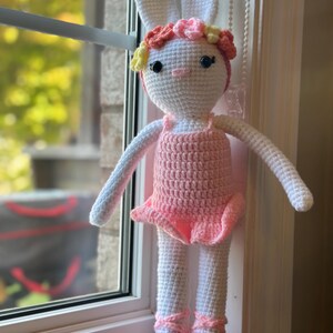 Puede incluir: Muñeca de conejo de ganchillo hecha a mano con un atuendo de bailarina. La muñeca es principalmente blanca con detalles en rosa, como un tutú, zapatillas de ballet y una diadema floral. El conejo tiene orejas largas y está cerca de una ventana.