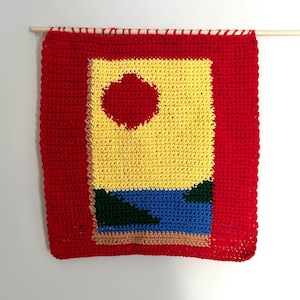 May include: A crocheted wall hanging featuring a geometric design in red, yellow, blue and green. The design includes a red circle in the centre of a yellow square, with a blue and green landscape below.