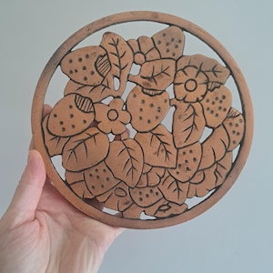 May include: A round, copper-colored trivet with a cut-out design of strawberries, leaves, and flowers. The trivet has a circular border and is held by a hand against a light background. The design is detailed and the trivet appears to be made of metal.