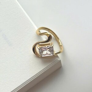 May include: A gold ear cuff featuring a rectangular, clear gemstone. The cuff has a curved, open design. The ear cuff is on a white surface next to a book. The book's title is partially visible.