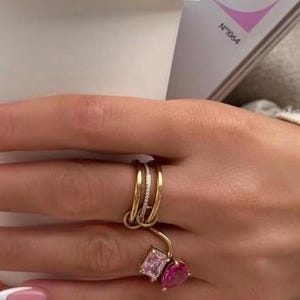 May include: A gold ring set featuring three bands, one with small clear stones and two with larger pink rectangular and pear-shaped gemstones. The rings are worn on a finger with a French manicure, holding a white paper coffee cup.
