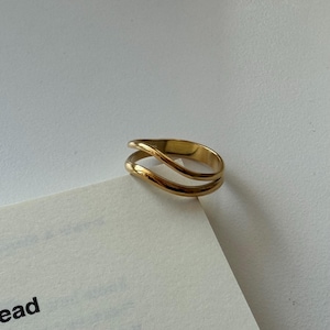 Könnte beinhalten: Ein goldfarbener Ring mit einem Doppelband-Design und einer glatten, polierten Oberfläche. Das Band besteht aus zwei parallelen, leicht geschwungenen Linien, die ein offenes, geschichtetes Erscheinungsbild erzeugen und eine reflektierende Oberfläche.