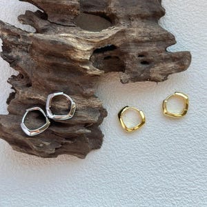 Puede incluir: Dos pares de pendientes de aro, uno plateado y otro dorado. Los pendientes tienen una forma geométrica, similar a un cuadrado. Los pendientes plateados están a la izquierda y los dorados a la derecha. Los pendientes se exhiben sobre un trozo de madera flotante.