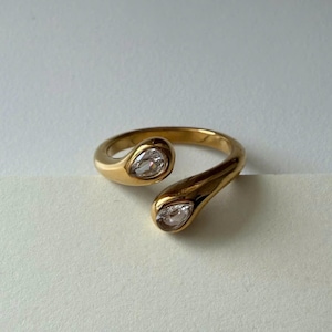 Peut inclure: Une bague dorée au design ouvert, ornée de deux pierres précieuses claires en forme de poire. La bague est en métal poli et se détache sur un fond blanc. Le design de la bague est moderne et élégant.