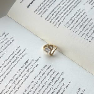 May include: A gold-toned ring featuring a single rectangular clear gemstone set within a curved, open-ended band. The ring rests on an open page of a book containing printed text about bread recipes, including ingredients like corn, wheat, and rye.