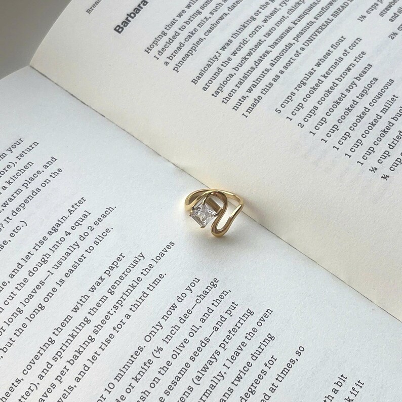 May include: A gold-toned ring featuring a single rectangular clear gemstone set within a curved, open-ended band. The ring rests on an open page of a book containing printed text about bread recipes, including ingredients like corn, wheat, and rye.