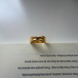 Pode incluir: Um anel dourado com um design único e entrelaçado. O anel tem um acabamento liso e polido e uma banda espessa. O anel está em cima de um livro com texto impresso.