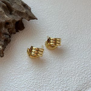May include: A pair of gold-colored hoop earrings. Each earring features a stacked, coiled design, creating a layered effect. The earrings are set against a textured, off-white surface, with a piece of driftwood in the background.
