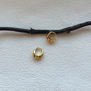 以下が含まれることがあります： 滑らかで丸みを帯びたデザインの、2つのゴールドカラーのフープピアス。イヤリングは、背景に濃い茶色の小枝がある、質感のある白い表面に表示されています。イヤリングは、研磨された反射面を持っています。