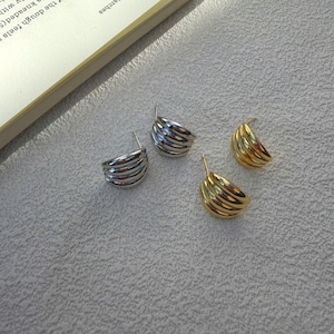 May include: Four earrings: two silver and two gold. Each earring has a curved, ridged design. The earrings are small, likely for pierced ears. The earrings are on a light grey surface.