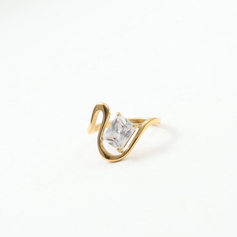 May include: A gold-toned ring featuring a single rectangular clear gemstone set within a curved, open-ended band. The metal band has a smooth, polished finish that wraps around the central stone, creating a fluid and asymmetrical design.