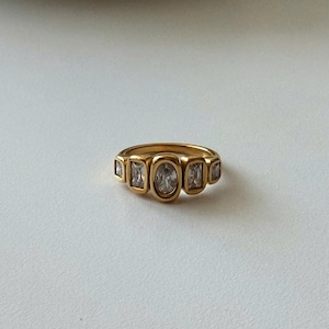 May include: A gold-toned ring featuring five clear, faceted stones set in a row. The centre stone is oval-shaped, flanked by two rectangular stones on each side, all encased in smooth, rounded metal bezels that create a continuous, polished band.