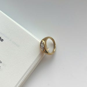 May include: A gold-toned ring featuring a smooth, polished band and a single marquise-cut clear gemstone set horizontally in the center. The ring has a thick, rounded profile and a reflective metallic finish that catches the light.