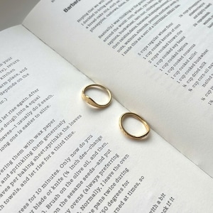 May include: Two gold rings with distinct designs are displayed on an open cookbook. One ring has a leaf-like motif, the other a textured pattern. The rings are set against a backdrop of printed text.