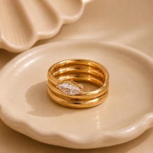 May include: A gold-toned ring featuring a stacked band design with three distinct layers. The top layer is adorned with a single marquise-cut clear gemstone set horizontally, creating a focal point on the polished metallic surface of the ring.