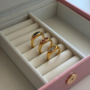 以下が含まれることがあります： ジュエリーボックスに入った、楕円形の宝石が付いた3つのゴールドリング。 宝石は緑、ピンク、白です。 リングは、ピンクのジュエリーボックス内の白いパッド入りの表面に表示されています。