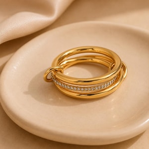 Puede incluir: Un anillo de varias bandas en tono dorado que presenta tres aros circulares interconectados. La banda central está incrustada con una hilera de pequeñas piedras claras y brillantes, mientras que las dos bandas exteriores son de metal liso y pulido.