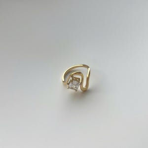 May include: A gold ring featuring a single rectangular clear gemstone set within a curved, open-ended band. The metal has a polished finish, and the stone is positioned centrally between the two ends of the wavy, fluid-shaped gold band.