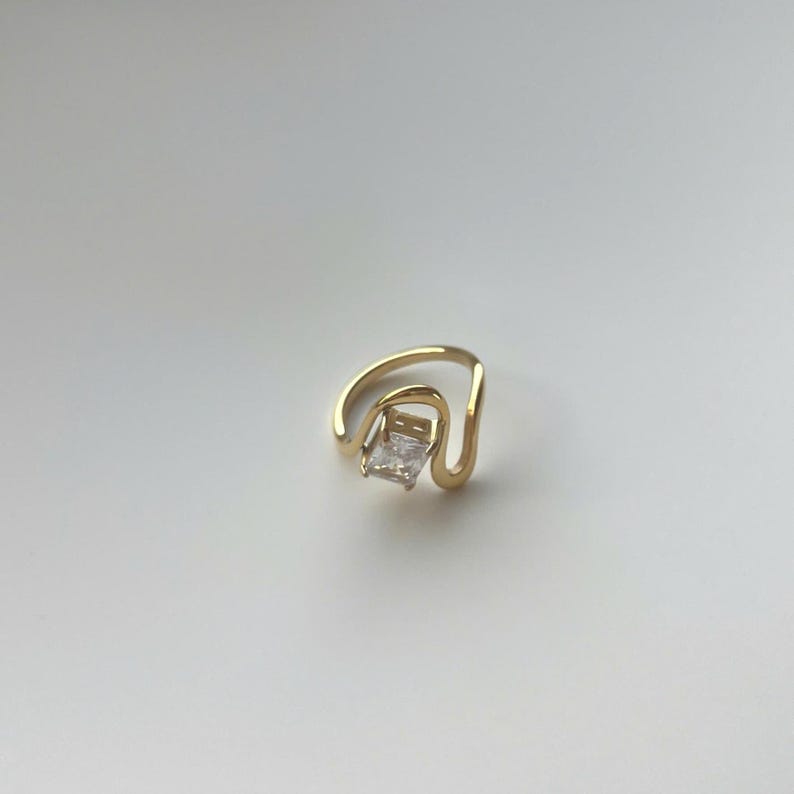 May include: A gold ring featuring a single rectangular clear gemstone set within a curved, open-ended band. The metal has a polished finish, and the stone is positioned centrally between the two ends of the wavy, fluid-shaped gold band.