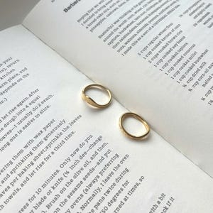 Può includere: Due anelli d'oro con design unici sono appoggiati su una pagina di libro di cucina aperta. Un anello presenta un design a forma di foglia, mentre l'altro ha un motivo strutturato. Gli anelli sono posti su uno sfondo di testo stampato.