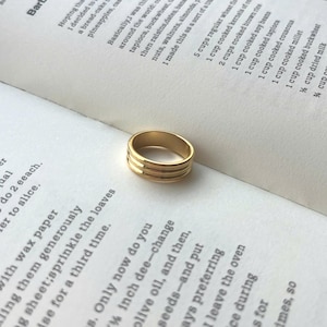 Peut inclure: Une bague dorée avec trois bandes horizontales, posée sur un livre ouvert avec du texte imprimé. Un bijou simple et élégant.