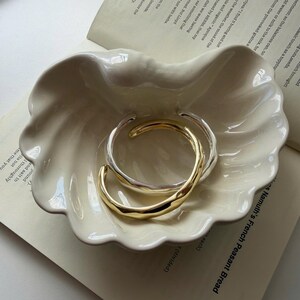 Puede incluir: Dos brazaletes abiertos, uno en tono dorado y otro en tono plateado, con una textura superficial lisa, irregular y ondulada. Los brazaletes están colocados juntos dentro de un plato de cerámica color crema con forma de concha.
