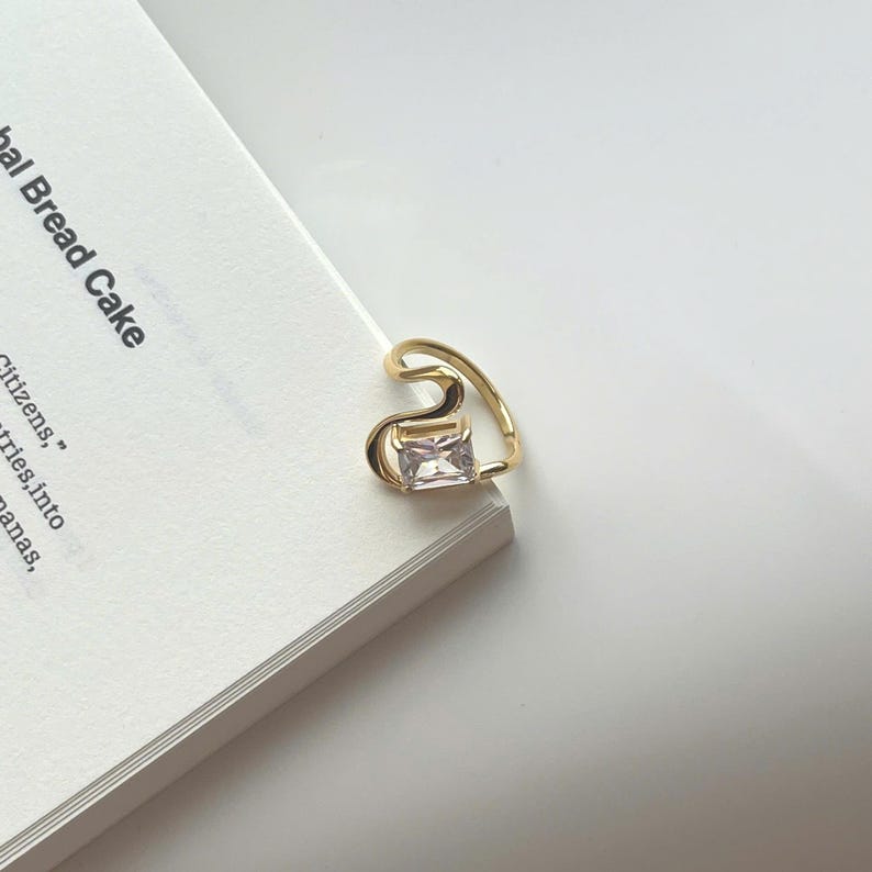 May include: A gold ear cuff featuring a rectangular, clear gemstone. The cuff has a curved, open design. The ear cuff is on a white surface next to a book. The book's title is partially visible.