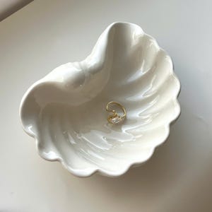 May include: A gold-toned ring featuring a single rectangular clear stone, resting inside a white ceramic dish shaped like a seashell. The dish has a glossy finish with curved, scalloped edges that mimic the natural ridges of a clam shell.