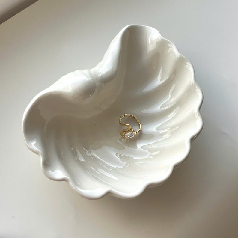 May include: A gold-toned ring featuring a single rectangular clear stone, resting inside a white ceramic dish shaped like a seashell. The dish has a glossy finish with curved, scalloped edges that mimic the natural ridges of a clam shell.
