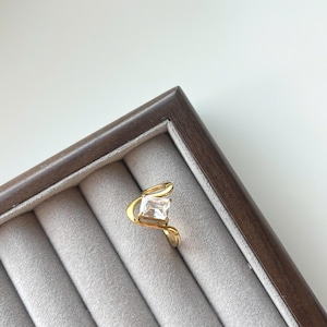 May include: A gold-toned ring featuring a single rectangular-cut clear gemstone set in a curved, open-style band. The metal band has a smooth, polished finish that wraps around the central stone, creating a distinct wave-like shape on the finger.