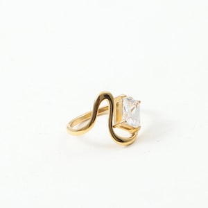 May include: A gold-toned ring featuring a smooth, undulating wave-shaped band that curves upward to hold a single, clear, rectangular-cut gemstone set in a simple metal frame. The design creates an open, asymmetrical silhouette on the finger.